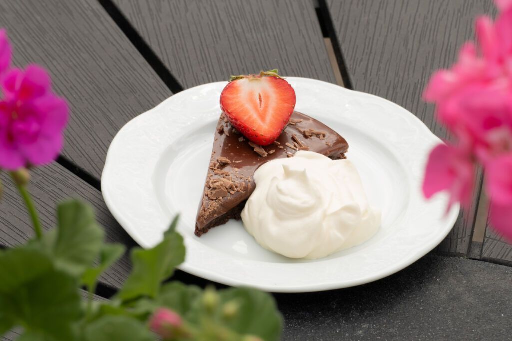 Bild på en chokladkaka med grädde och en jordgubbe på. Fatet står på ett bord och runt om syns blommor